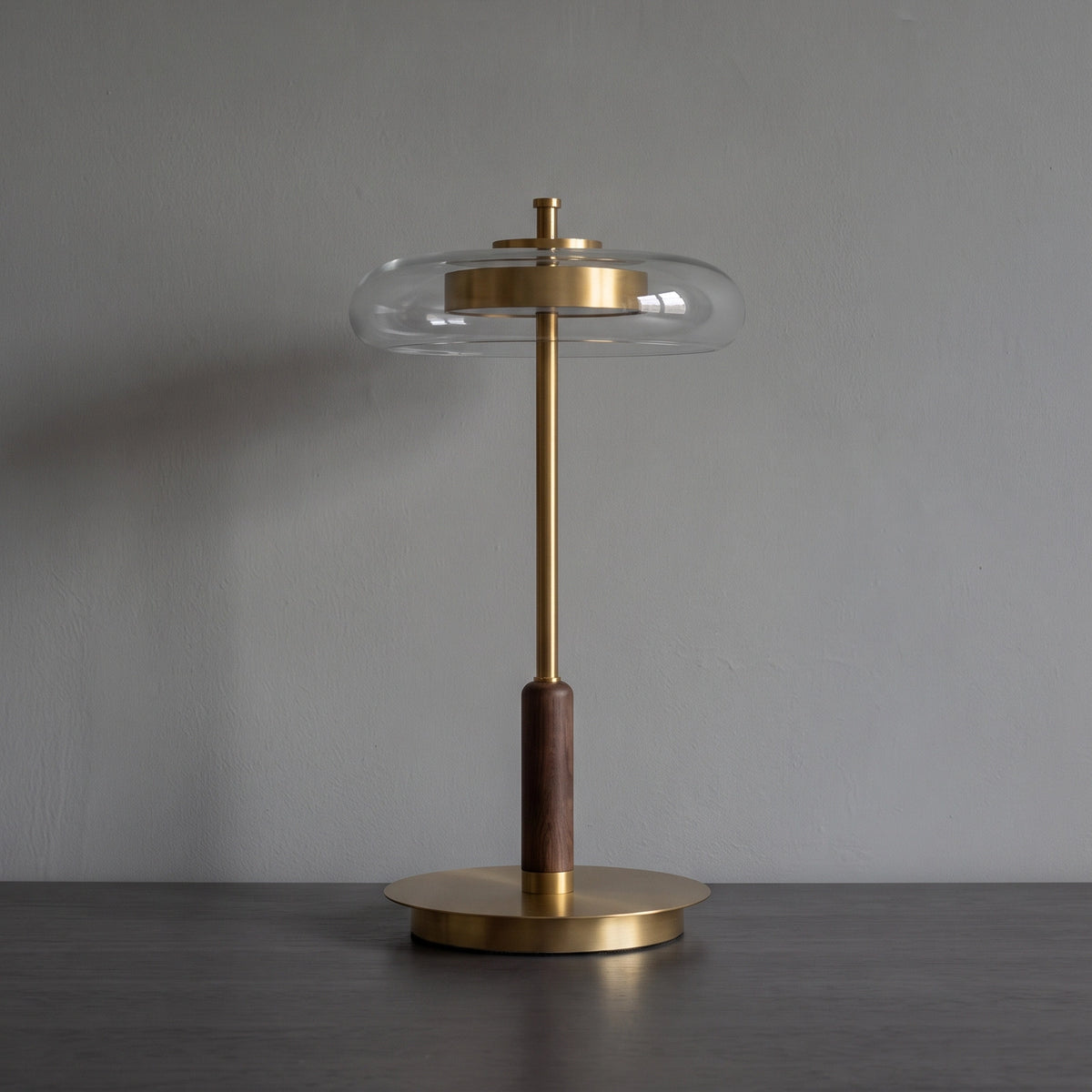 The Loria lamp switched off against a concrete wall, showing the clear glass halo, brass drum, walnut grip and round base.