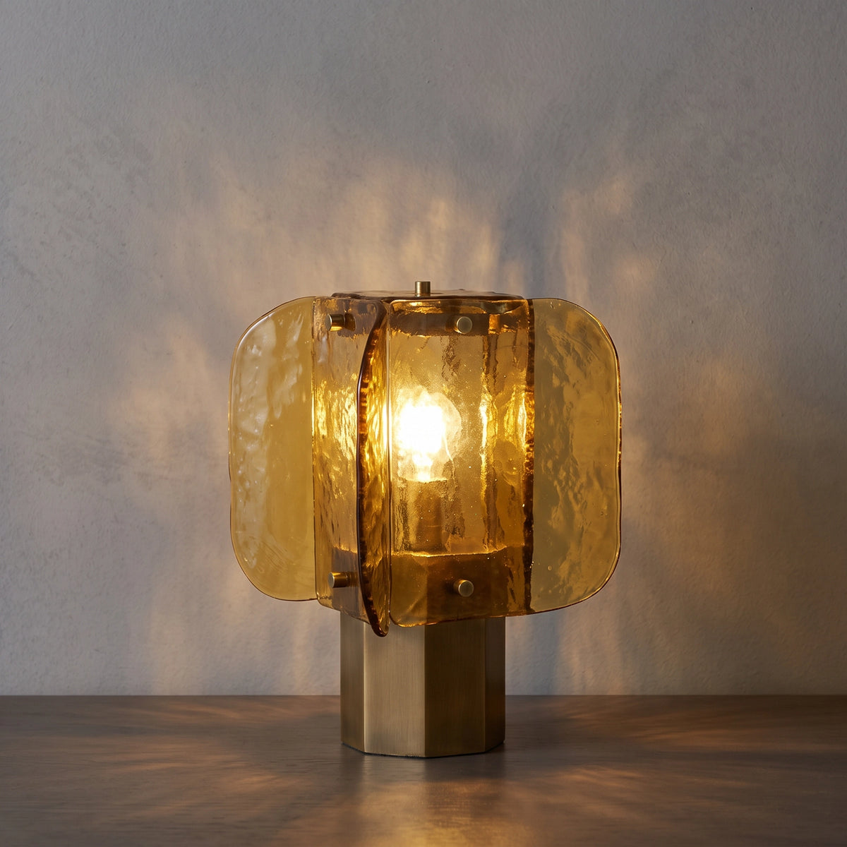 The Tramonto amber glass table lamp switched on, casting warm wave-like light patterns on the wall and a dark wood table.